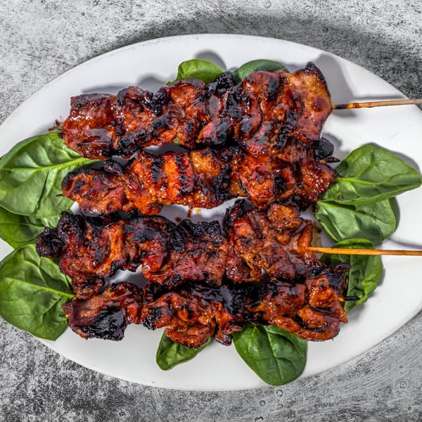 Three skewers of glistening, dark Filipino pork barbecue on a white plate with spinach leaves.