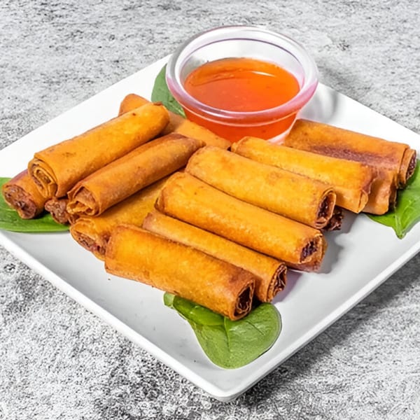 A white plate filled with golden-brown Filipino lumpia rolls, garnished with spinach and a side of sweet chili sauce.