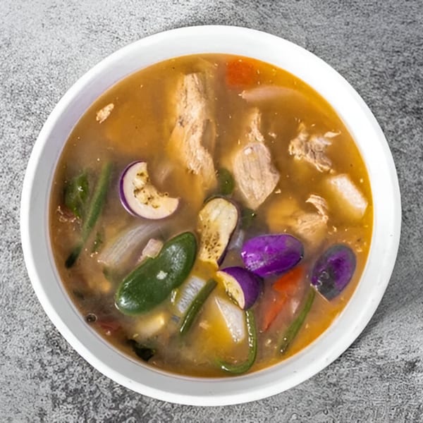 Overhead view of a Filipino soup with meat, purple and white eggplant, and other vegetables in a white bowl.
